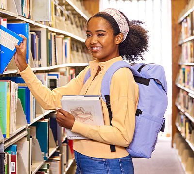 Girl wearing a backpack grabbing a book in the library.