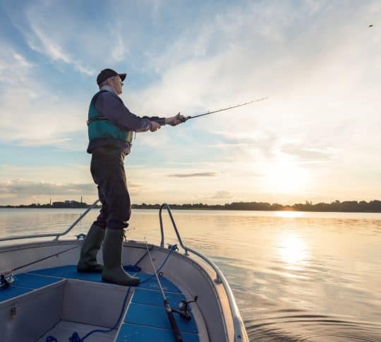 Man standing on the edge of a boat casting a fishing rod into a lake