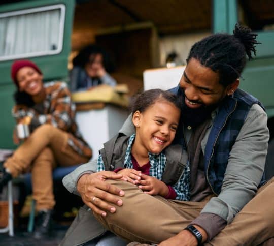 Man hugging daughter with camper and family in the background